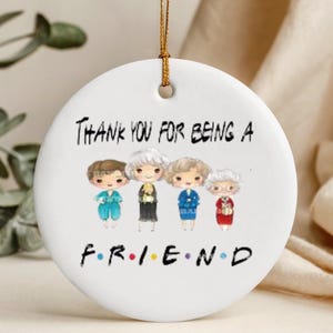 May include: A white ceramic ornament with a gold string for hanging. The ornament features a cartoon illustration of four women and the text "Thank you for being a friend" with the word "FRIEND" in the style of the TV show "Friends".