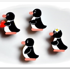 Könnte beinhalten: Vier bemalte Holz-Pinguin-Figuren in Schwarz und Weiß mit rosa Akzenten. Ein Pinguin hält ein rotes Herz, ein anderer hat eine weiße Blume auf dem Kopf.