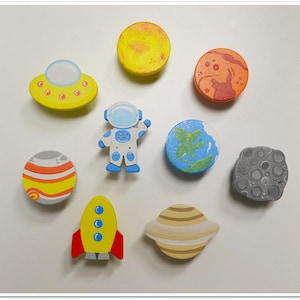 May include: A collection of colorful wooden space-themed magnets. Includes a yellow UFO, astronaut, rocket, planets, sun, moon, and Saturn. The magnets are painted with bright colors and are likely for decorative or educational purposes.