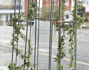 Op de afbeelding: Drie metalen tuinbogen met een witte afwerking, versierd met kunstgras en roze bloemen. De bogen staan op een bed van kunstgras. Een bordje geeft aan "£20 each,  vanised off".
