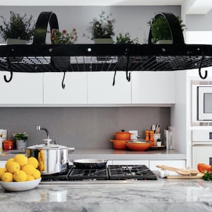 Puede incluir: Un estante para ollas de metal negro con ganchos que cuelgan del techo sobre una encimera de cocina. El estante es de forma ovalada y tiene un diseño de malla de alambre. Hay ollas y sartenes colgando de los ganchos.