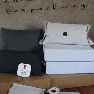 May include: Sustainable packaging with a white box, a brown box, and a gray pillow. The pillow has a sticker that says "M&auml;dchen sucht Kleid". There are also black and gold "Thank You" stickers.