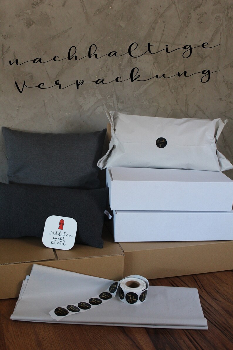 May include: Sustainable packaging with a white box, a brown box, and a gray pillow. The pillow has a sticker that says "M&auml;dchen sucht Kleid". There are also black and gold "Thank You" stickers.
