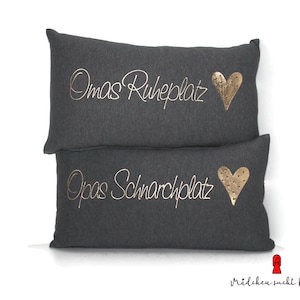Può includere: Due federe per cuscini grigie con lettere dorate e cuori. La federa superiore recita "Oma's Ruheplatz" e la federa inferiore recita "Opa's Schnarplatz".