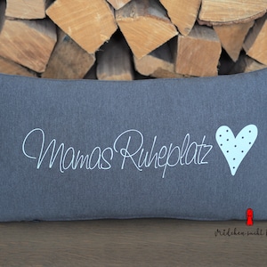 Op de afbeelding: Grijs kussen met witte geborduurde tekst "Mamas Ruheplatz" en een lichtblauw hart met witte stippen.