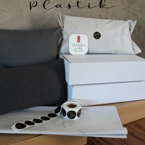 May include: A brown cardboard box with white tissue paper and black and gold thank you stickers. Two gray pillows are on top of the box. A white box with a red and white sticker that says "M&auml;dchen sucht Kleid" is on top of the pillows. The background is a gray wall with the text "Verpackung ohne Plastik" in black.