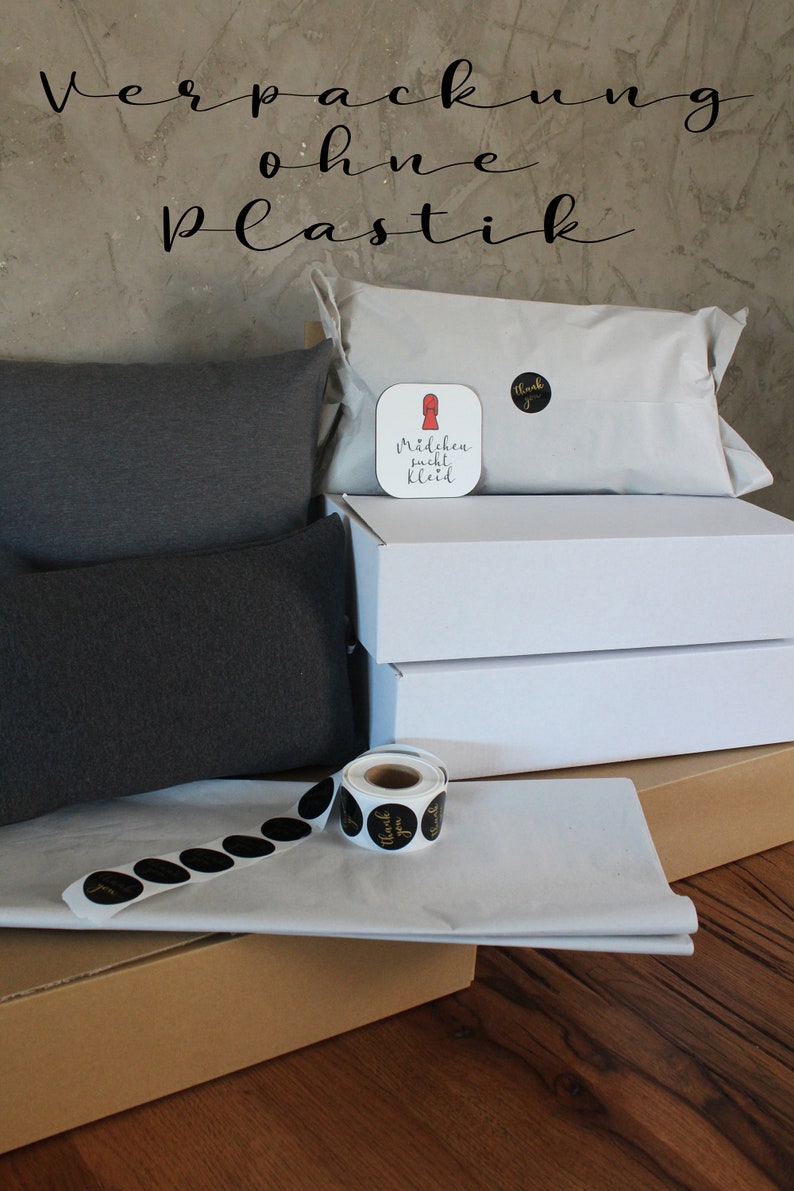 May include: A brown cardboard box with white tissue paper and black and gold thank you stickers. Two gray pillows are on top of the box. A white box with a red and white sticker that says "M&auml;dchen sucht Kleid" is on top of the pillows. The background is a gray wall with the text "Verpackung ohne Plastik" in black.