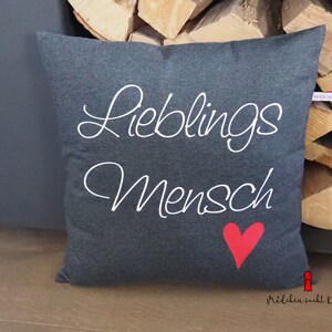 Könnte beinhalten: Graues Kissen mit weißem Text "Lieblings Mensch" und einem roten Herz. Das Kissen liegt auf einer Holzoberfläche.