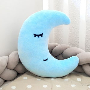 Könnte beinhalten: Ein hellblaues, sichelförmiges Kissen in Mondform mit schwarzen gestickten Augen und einem Lächeln. Das Kissen liegt auf einem geflochtenen grauen Kissen.