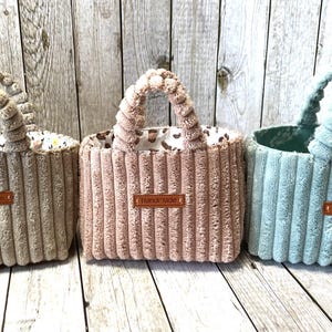 Könnte beinhalten: Drei handgefertigte Cord-Tragetaschen in Beige, Rosa und Blau. Jede Tasche hat einen passenden Griff und ein braunes "handmade"-Etikett. Die beige Tasche hat ein Blumenfutter, die rosa Tasche ein Leopardenmuster und die blaue Tasche ein gepunktetes Futter.