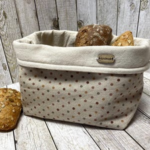 Könnte beinhalten: Ein beiger Brotbeutel mit braunen Punkten, der zwei Brote enthält. Der Korb hat ein cremefarbenes Futter und ein kleines "Handmade"-Etikett. Ein Brot liegt links vom Korb.