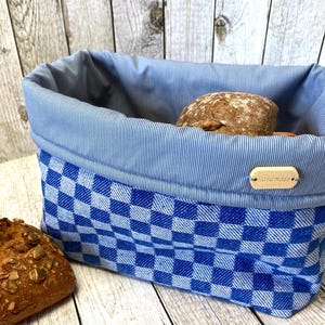 Könnte beinhalten: Ein blau-weiß karierter Brotkorb mit blau gestreiftem Innenfutter. Der Korb enthält zwei Brotlaibe. Ein kleines Holzschild mit der Aufschrift "Hand made" ist am Korb befestigt.
