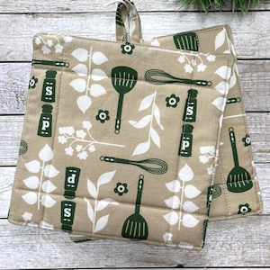Könnte beinhalten: Zwei beigefarbene Topflappen mit einem grün-weißen Muster. Das Muster enthält Blätter, Blumen, Schneebesen, Spatel und Salz- und Pfefferstreuer. Die Topflappen haben eine Schlaufe zum Aufhängen.