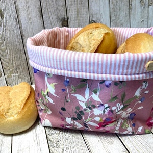 Könnte beinhalten: Ein rosa Stoff-Brotkorb mit weißen Streifen und einem Blumenmuster. Der Korb ist mit drei weißen Brötchen gefüllt.