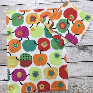 Könnte beinhalten: Ein weißes Küchentuch und Topflappen-Set mit einem bunten Apfelmuster. Die Äpfel sind in Rot, Grün, Orange und Gelb gehalten, mit schwarzen Stielen und floralen Details. Das Tuch hat eine weiße Aufhängeschlaufe.