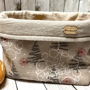 Könnte beinhalten: Ein beigefarbener Stoffbrotkorb mit einem braunen und weißen Weihnachtsbaum-Muster und dem Text "Frohe Weihnachten" in Kursivschrift. Der Korb hat ein braunes Leder-Etikett mit der Aufschrift "Handmade".