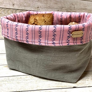 Könnte beinhalten: Ein grauer Leinen-Brotkorb mit einer rosa-blauen, floralen Futter. Der Korb ist mit Brötchen gefüllt und hat ein "Handmade"-Etikett am Futter befestigt.
