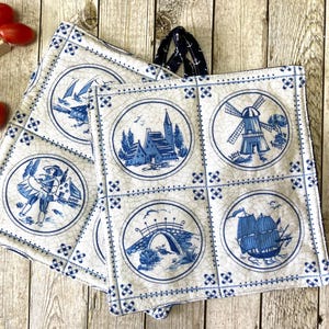 Könnte beinhalten: Zwei weiß-blaue Ofenhandschuhe im niederländischen Stil mit einem marineblauen Griff. Jeder Handschuh zeigt ein sich wiederholendes Muster aus kreisförmigen Illustrationen, die Windmühlen, Schiffe und Brücken darstellen. Die Handschuhe sind aus gestepptem Stoff.