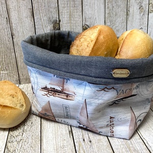 Könnte beinhalten: Ein grauer Stoff-Brotkorb mit einem weißen und braunen nautischen Druck. Der Korb hat einen grauen Denim-Oberteil mit einem braunen "Handmade"-Etikett. Der Korb ist mit vier weißen Brötchen gefüllt.