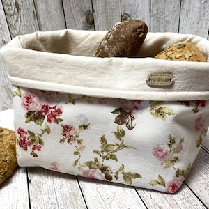 Könnte beinhalten: Ein weißer Stoff-Brotkorb mit einem Blumenmuster mit rosa Rosen und grünen Blättern. Der Korb hat einen cremefarbenen oberen Rand mit dem Wort "handgefertigt" auf einem braunen Etikett. Der Korb ist mit verschiedenen Broten gefüllt.