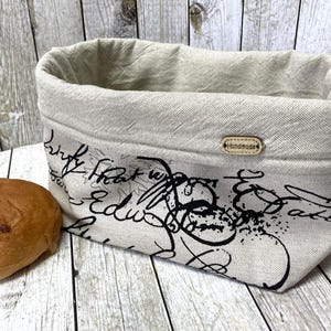 Könnte beinhalten: Ein weißer Brotkorb aus Leinen mit schwarzer Schrift und einem "Handmade"-Etikett. Der Korb ist mit einem runden, braunen Brotlaib gefüllt.