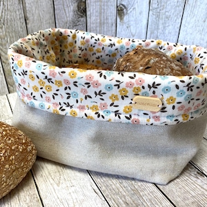 Könnte beinhalten: Ein Brotbeutel aus Leinen mit einem floralen Futter in Rosa, Gelb und Blau. Der Korb enthält ein Brot. Ein kleines Holzschild mit dem Wort "handmade" ist am Korb befestigt.