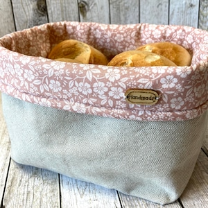 Könnte beinhalten: Ein Brotkorb aus rosa-weißem Blumenstoff mit einer grauen Leinenfutter. Der Korb hat ein Holzschild mit der Aufschrift "Handmade".