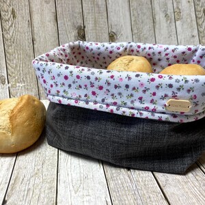 Könnte beinhalten: Ein grauer und weißer Stoff-Brotkorb mit Blumenmuster und einem Holzschild mit der Aufschrift "Hand Made". Der Korb ist mit vier weißen Brötchen gefüllt.
