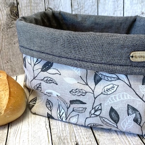 Könnte beinhalten: Ein rechteckiger Stoffkorb mit beiger Außenseite, einem schwarz-weißen Blattmuster und einem dunkelgrauen, umgeschlagenen Rand in Denim-Optik. Ein kleines ovales Holzschild mit der Aufschrift Handmade ist vorne am dunkelgrauen Rand befestigt.
