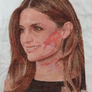 May include: Cross-stitch portrait of a woman with long brown hair, wearing a black shirt with a floral pattern. The woman is smiling and looking to the side.