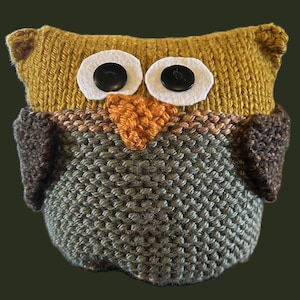 May include: A knitted owl toy made with brown, green and yellow yarn. The owl has large white eyes with black pupils and a small orange beak.
