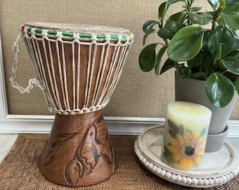 May include: A brown wooden djembe drum with a white rope design and a green, red, and yellow trim around the top. The drum is sitting on a brown woven mat.