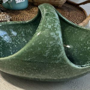 Könnte beinhalten: Ein grüner Keramikkorb mit Henkel. Der Korb hat ein gesprenkeltes Muster und ein geschwungenes Design. Der Griff ist gewölbt und der Korb hat zwei offene Fächer. Der Korb ist wahrscheinlich Vintage und für dekorative Zwecke geeignet.