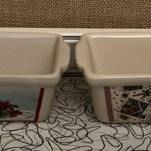 May include: Two rectangular ceramic loaf pans. One pan features a winter scene with a cardinal and poinsettias. The other pan has a pattern of snowflakes and holiday images. Both pans have a beige exterior.