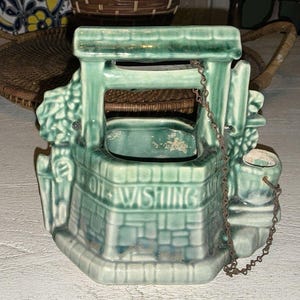 May include: A vintage ceramic wishing well planter in a teal green color. The planter has a brick-like design with the words "OH WISHING WELL" on the front. A small bucket hangs from a chain on the side.
