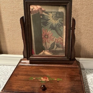May include: A vintage wooden vanity mirror with a rectangular frame and a small drawer. The mirror is framed in dark brown wood and has a decorative top. The drawer is adorned with a painted floral design and a small, round knob.
