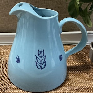 Könnte beinhalten: Eine hellblaue Keramikkrug mit gebogenem Griff und Ausguss. Die Krug hat ein Tulpenmuster in Dunkelblau. Der Krug wird wahrscheinlich zum Servieren von Getränken verwendet und hat ein klassisches, elegantes Design.