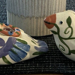 Könnte beinhalten: Zwei Keramik-Vogelfiguren mit cremefarbenen Körpern, orangefarbenen Schnäbeln, blauen Augen und dekorativen lila, grünen und blauen Blumenmustern. Die Vögel stehen nebeneinander, was auf ein dekoratives oder künstlerisches Thema hindeutet.