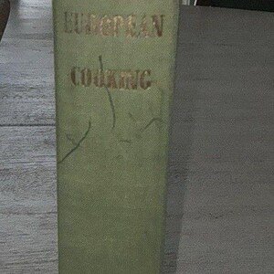 May include: A vintage cookbook titled "European Cooking" in a light green cover. The title is in gold lettering. The book is standing upright on a light-colored surface.