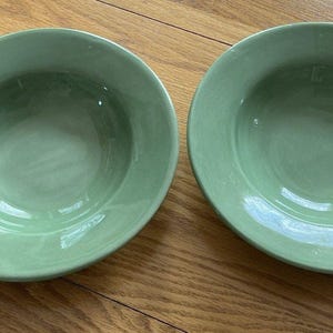 May include: Two pale green ceramic bowls with a wide rim. The bowls are round and have a smooth, glossy finish. The bowls are on a wooden surface.