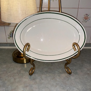 May include: An oval-shaped white ceramic serving platter featuring a thin, dark green band encircling the rim. The platter is displayed on a gold-toned metal stand with curved supports that hold the dish in an upright position.