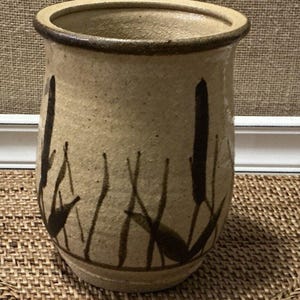 Puede incluir: Un jarrón de cerámica beige con forma cilíndrica. El jarrón presenta un borde marrón oscuro y está decorado con un diseño estilizado de espadañas en marrón oscuro. El jarrón está sobre una superficie tejida.