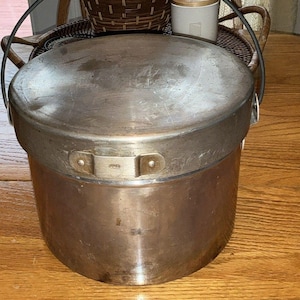May include: A vintage metal cooking pot with a lid and a handle. The pot is cylindrical with a slightly rounded base and a lid that fits snugly on top. The metal has a worn, aged appearance.