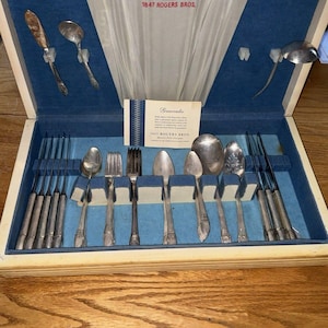 May include: A silver-plated flatware set in a wooden storage case with a blue velvet interior. The set includes various spoons, forks, and knives, with a central card labeled 1847 Rogers Bros. The case lid features a white fabric panel with the 1847 Rogers Bros logo.