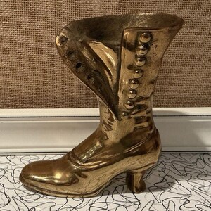 May include: A gold-colored, vintage-style boot sculpture. The boot features a high heel, button details, and a curved top. The sculpture is made of a shiny, metallic material and is likely a decorative item.