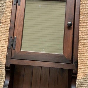 May include: A dark brown wooden wall cabinet featuring a single hinged door with a rectangular mirror insert, two black metal hinges, and a small round black knob. Below the door is an open storage shelf with a vertical plank back panel.
