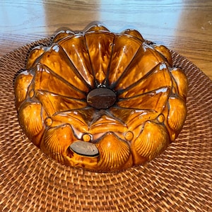 May include: A brown ceramic bundt pan with a decorative shell design. The pan has a central hole and is placed on a woven wicker mat. The pan's exterior is glazed with a glossy finish.