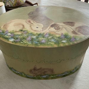Puede incluir: Una sombrerera redonda de estilo vintage con una escena pintada de tres conejos en un campo de flores. La caja es principalmente verde con un borde floral decorativo y un conejo marrón pintado en el lateral.