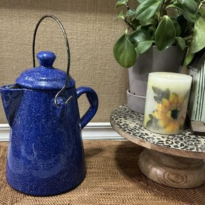 Puede incluir: Tetera de cerámica azul moteada con asa de metal y tapa. La tetera está sobre una estera tejida marrón.