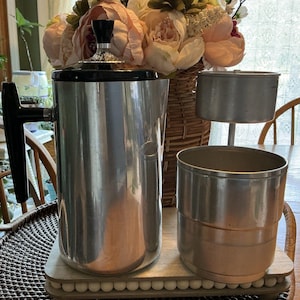 Puede incluir: Una cafetera percoladora de metal plateado con asa negra y tapa negra que presenta un pomo de cristal transparente. Junto a la cafetera hay una cesta de café cilíndrica de metal y un componente de base metálico a juego para el sistema.